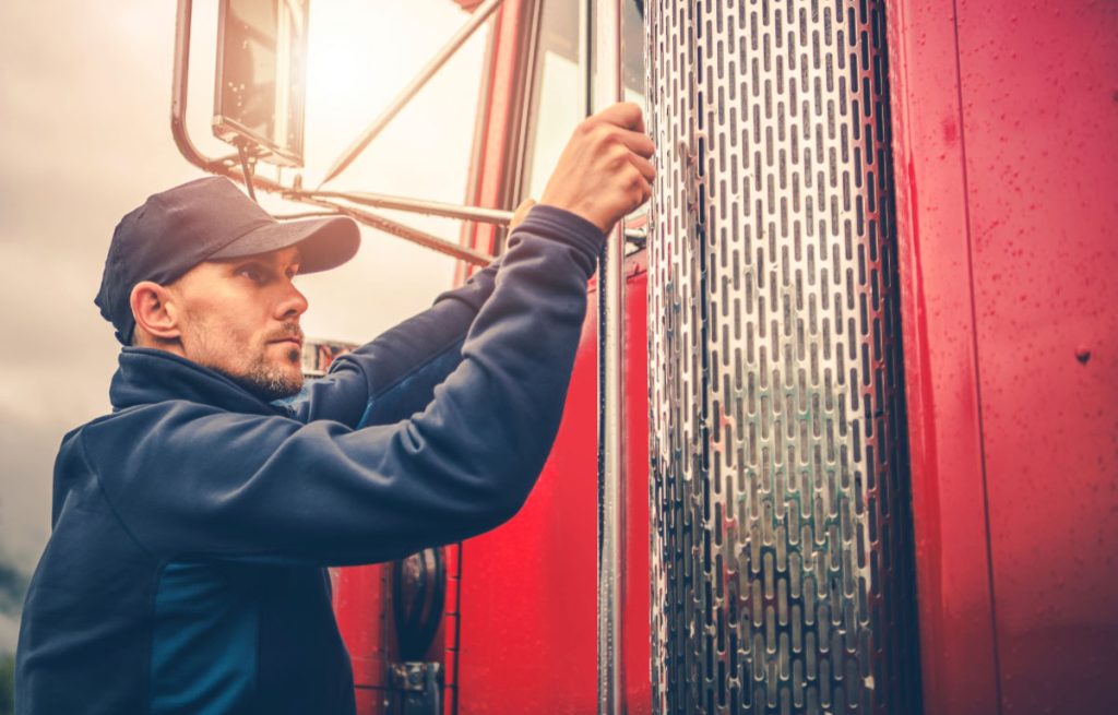 Commercial truck driver in a blue hoodie and cap climbing into the cab of a red semi-truck, representing regulated CMV driver operations under FMCSA requirements